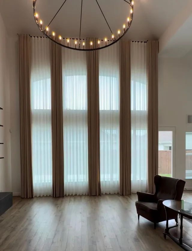Full-height beige curtains and white sheers framing arched windows beneath a ring chandelier.