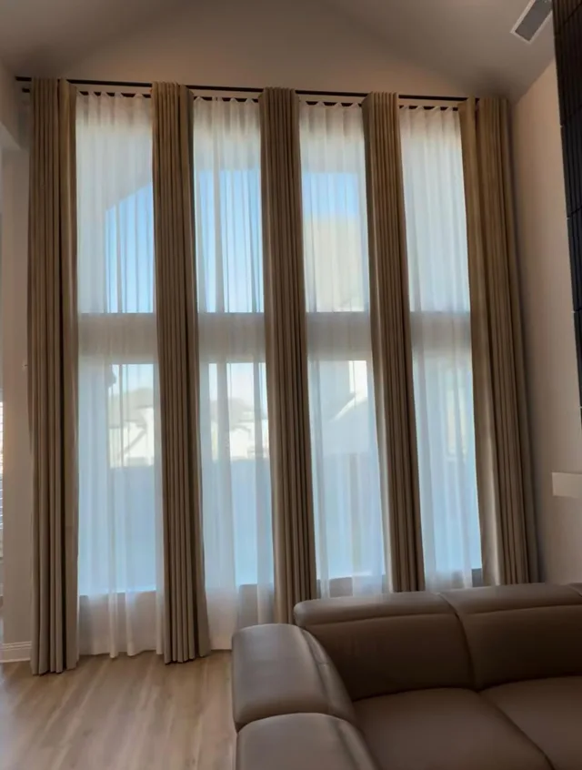 Side view of beige drapery and white sheers along a double-height living room window wall.