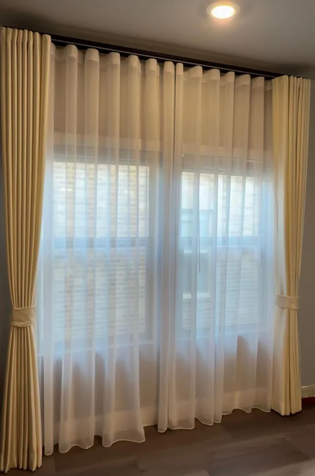 Pleated ivory side curtains with white sheer panels layered over horizontal blinds.